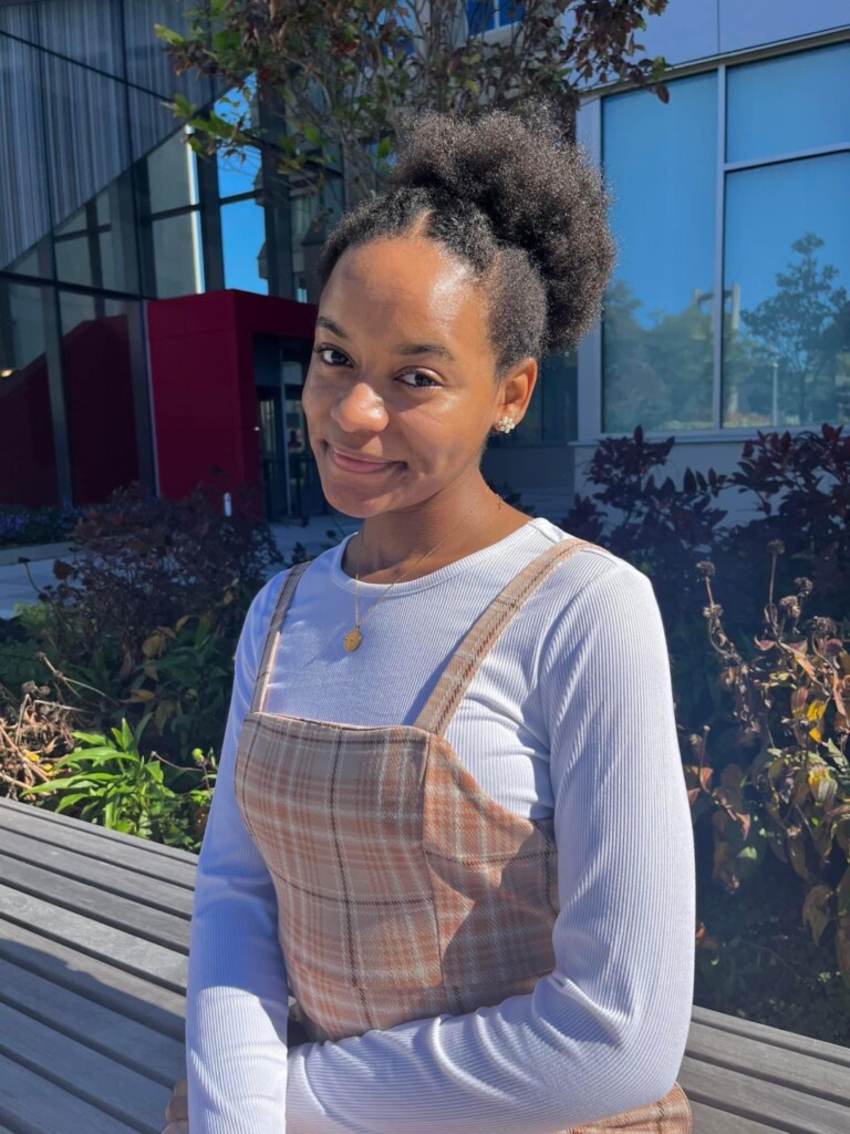 African American young woman with natural hair wearing a white shirt and plaid overalls.